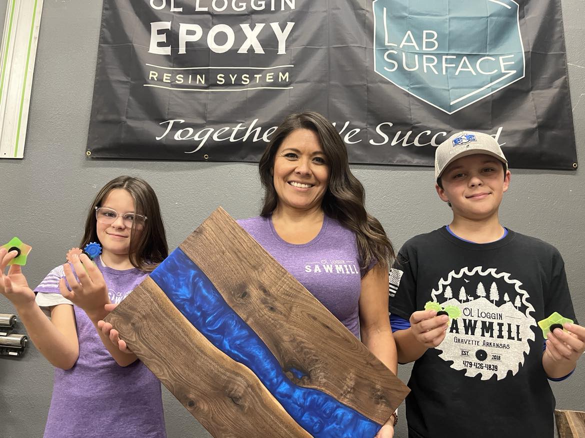 A woman holding a blue epoxy river serving tray. Two kids (girl left, boy right) are standing beside her holding up epoxy fidget spinners.